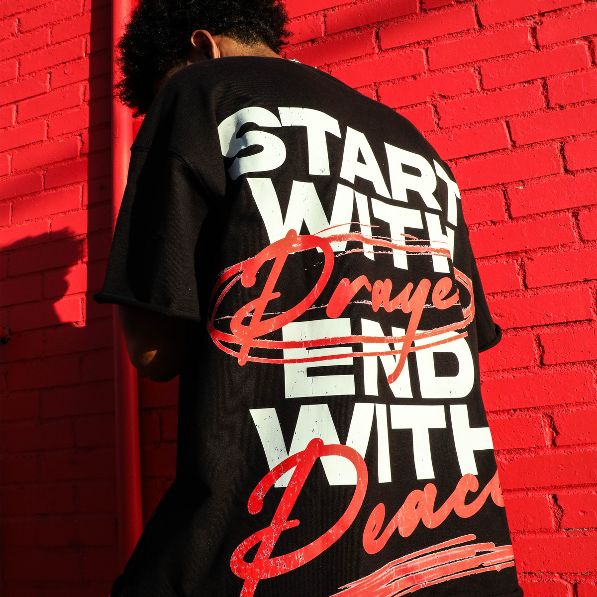 Back of the Saint Heights Start With Prayer tee worn against a red brick wall, oversized white type reading start with prayer end with peace with red script lettering across it.